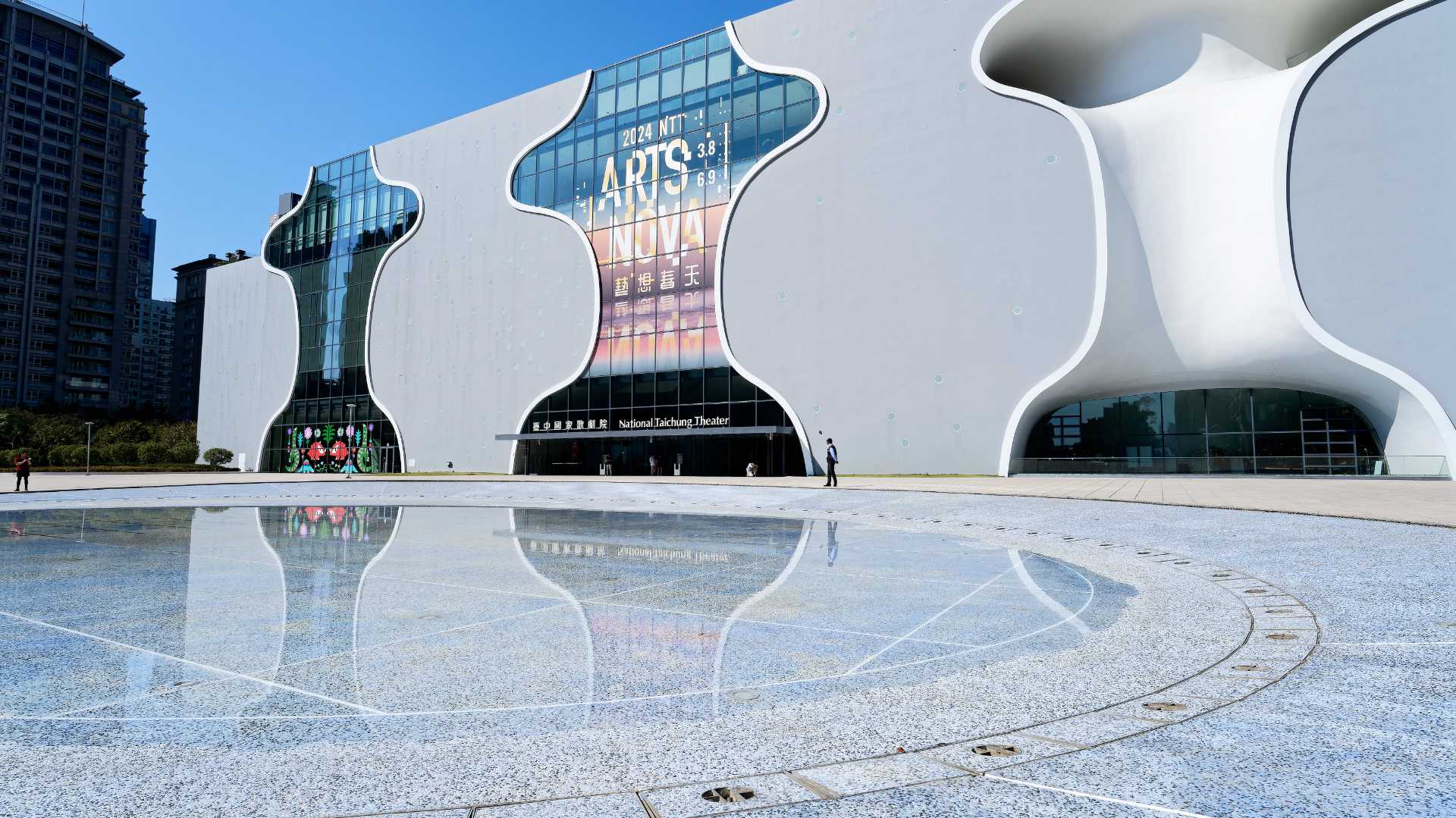 The National Taichung Theater building reflected in a shallow pool of water. The building is fundamentally rectangular, but punctuated by multi-level curved windows and concave balcony spaces.