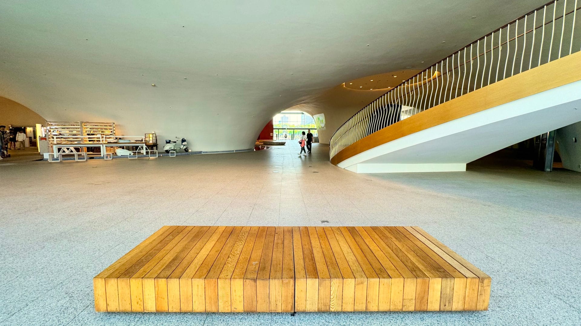 A low, rectangular wooden bench seat in the foreground. The vast, cuvaceous foyer of the National Taichung Theater is visible beyond.