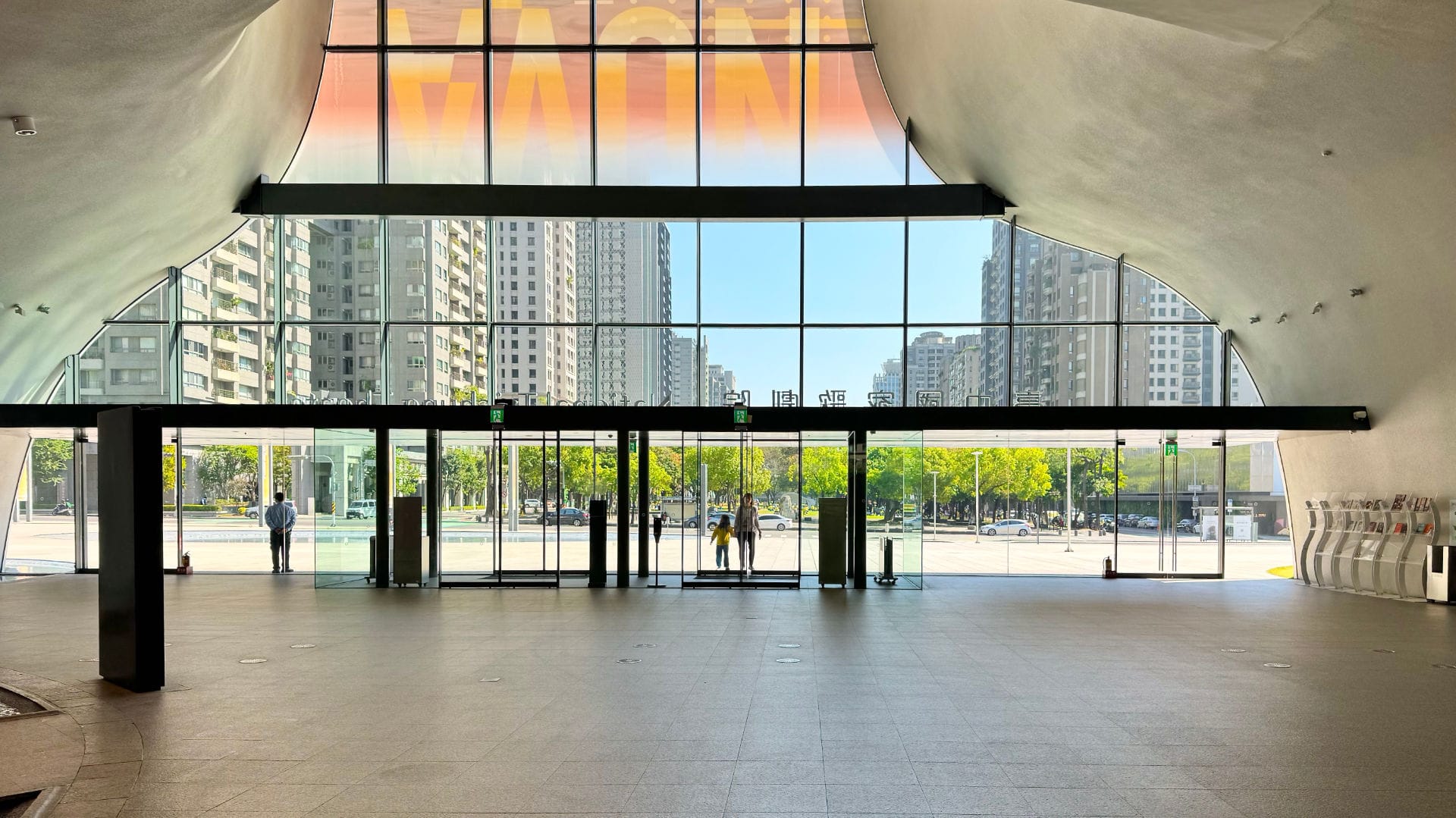 The triple-height foyer of National Taichung Theater. There is no one inside, but a parent and young child are entering through the glass doors. Through the glass windows and doors, Taichung city looks beautiful on a sunny day.