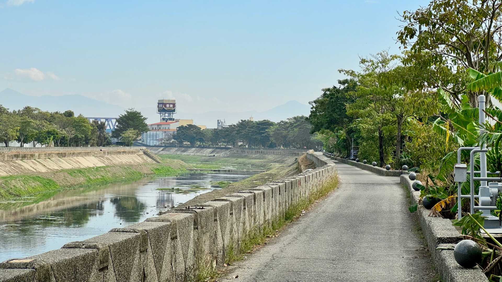 A narrow riverside road (one car-width wide). The river is unattractive, with concrete walls on either side. The area around the river is quite green with trees, including a banana tree in the foreground.