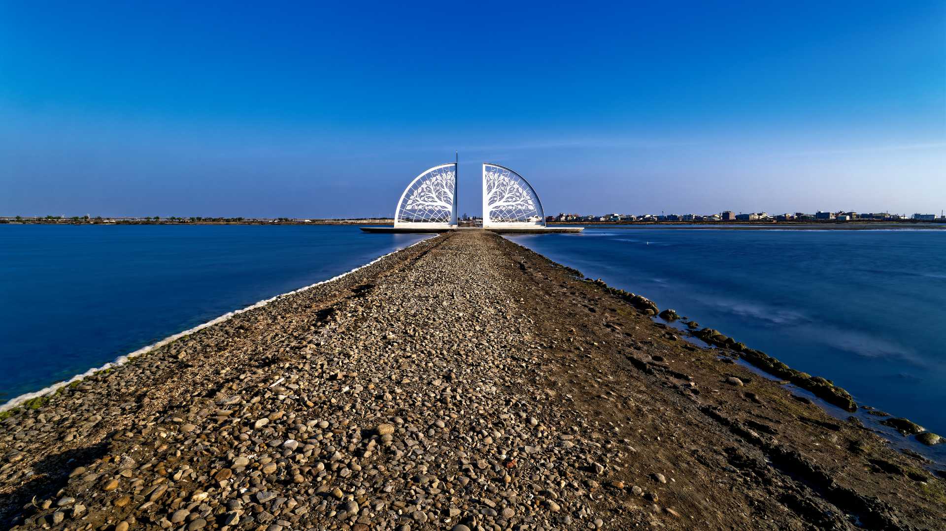 The Tree of Life sculpture on the Tainan salt flats. The sculpture looks like a very large fan, split in the middle to allow people to walk through. The fan has cut-out shapes that give it the appearance of a tree. It is far from the camera, but perhaps 10 or 15 meters tall and twice as wide.