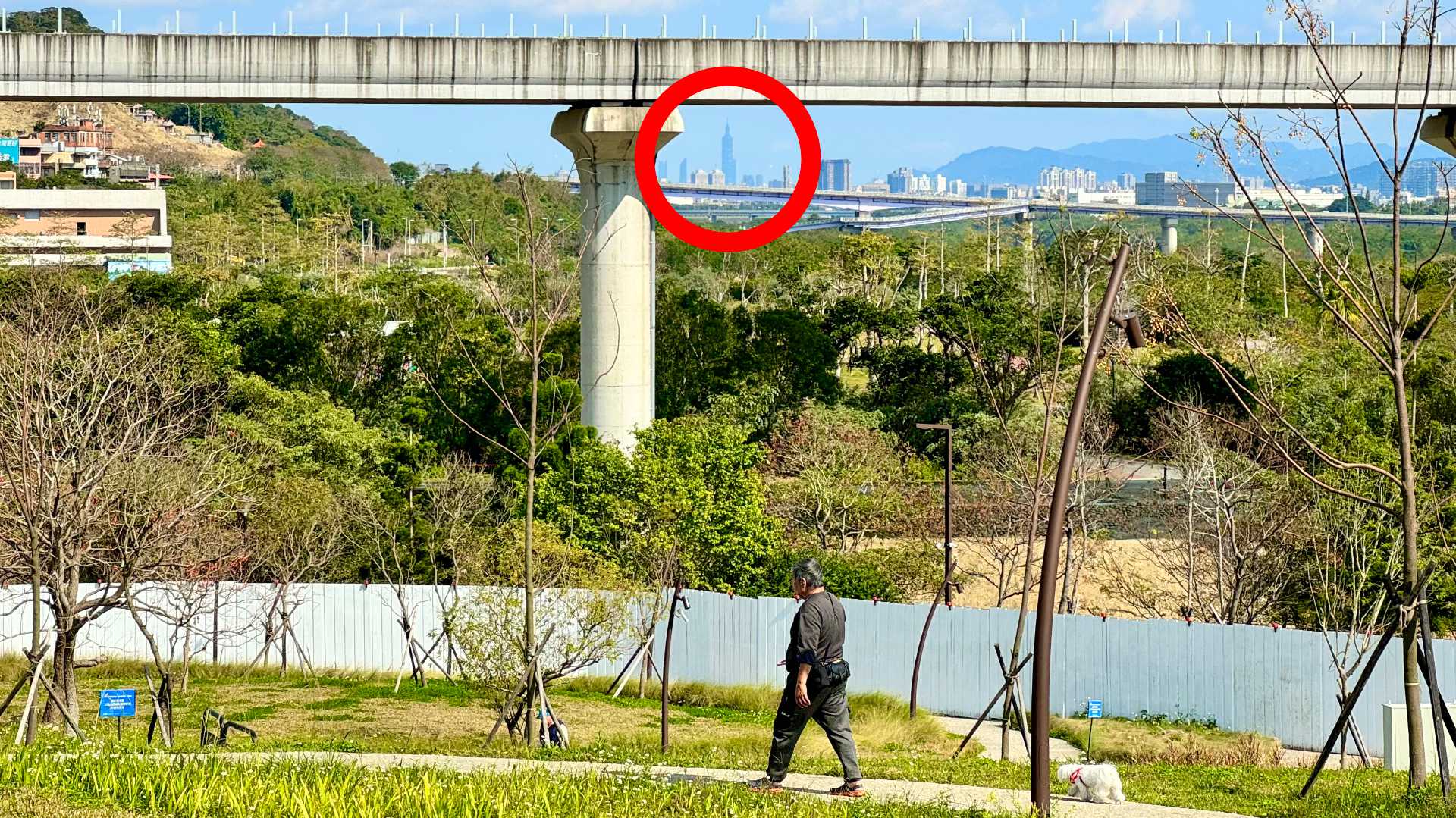 Photo looking across a public park and beneath an overbridge to the skyline of Taipei City in the distance. Taipei 101 is circled in the image.