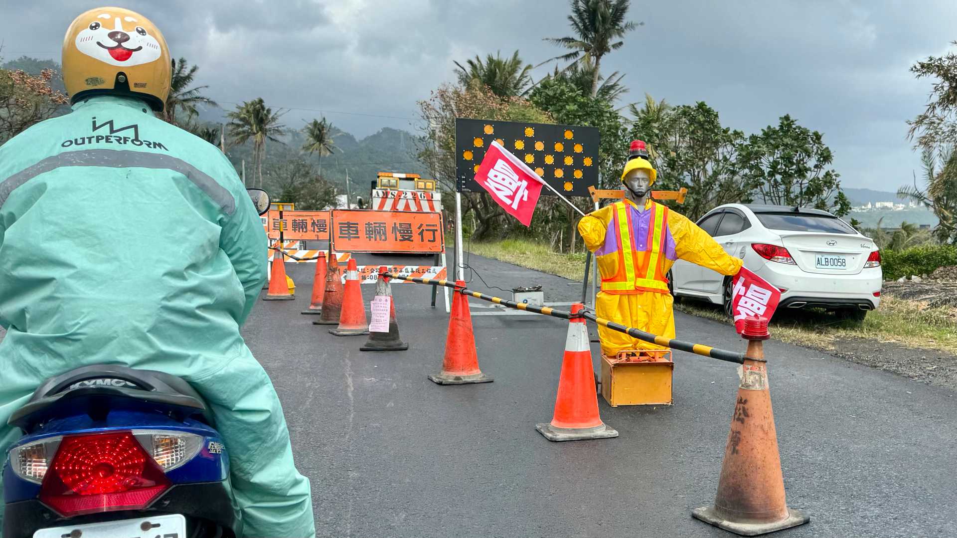 A mannequin in high-visibility clothing, in front of some roadworks, holding two flags that read ‘slow’ in Chinese.