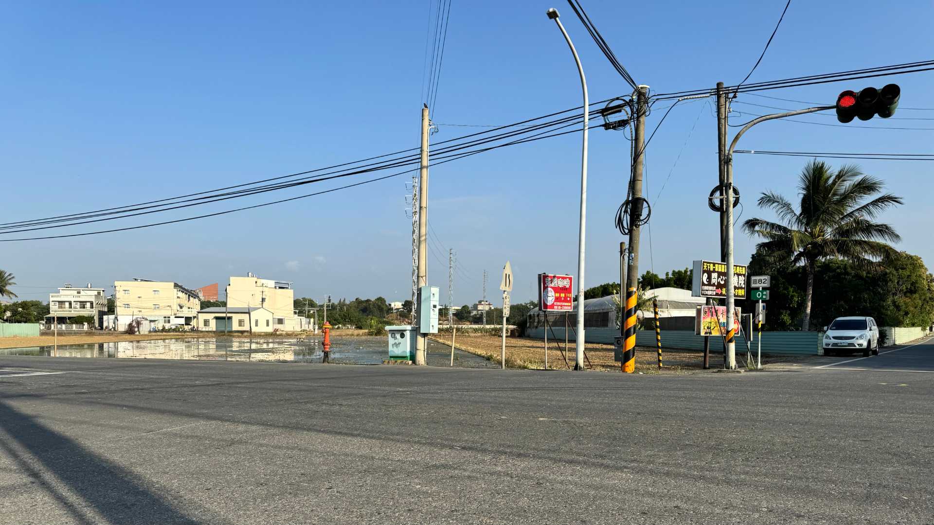 A country intersection with traffic lights. A paddy field is visible on the other side of the road, as well as a palm tree and some houses, with more trees in the distance.