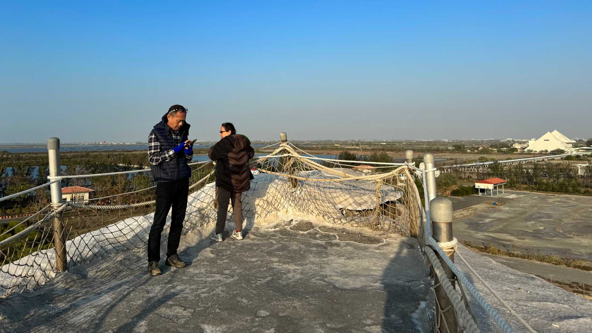 View from the top of Qigu Salt Mountain. There are two people standing on the top. The ground is a dirty gray where people have stood, but the rest of the salt mountain is white. The Tainan salt flats stretch out in the distance.