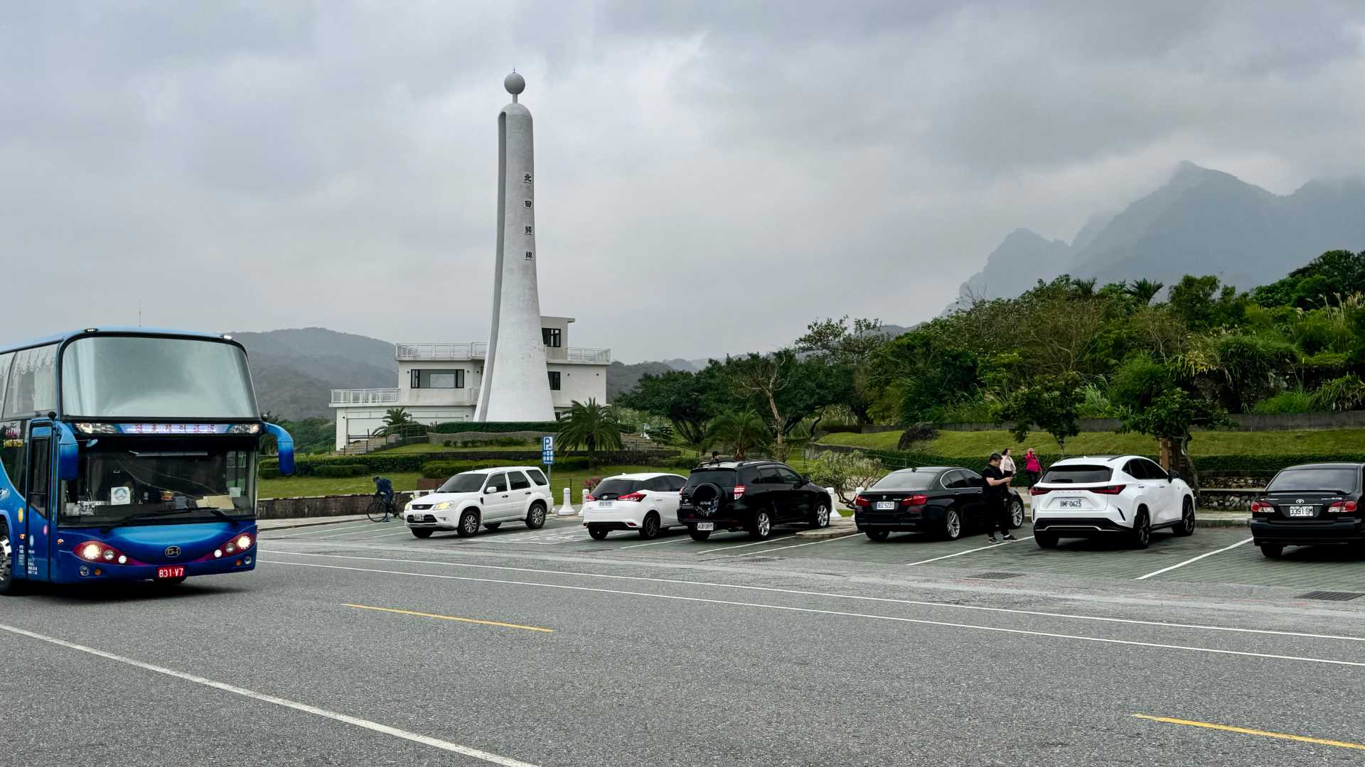 A tall, round sculpture with a ball on top, next to a busy carpark. A bus drives past in the foreground.