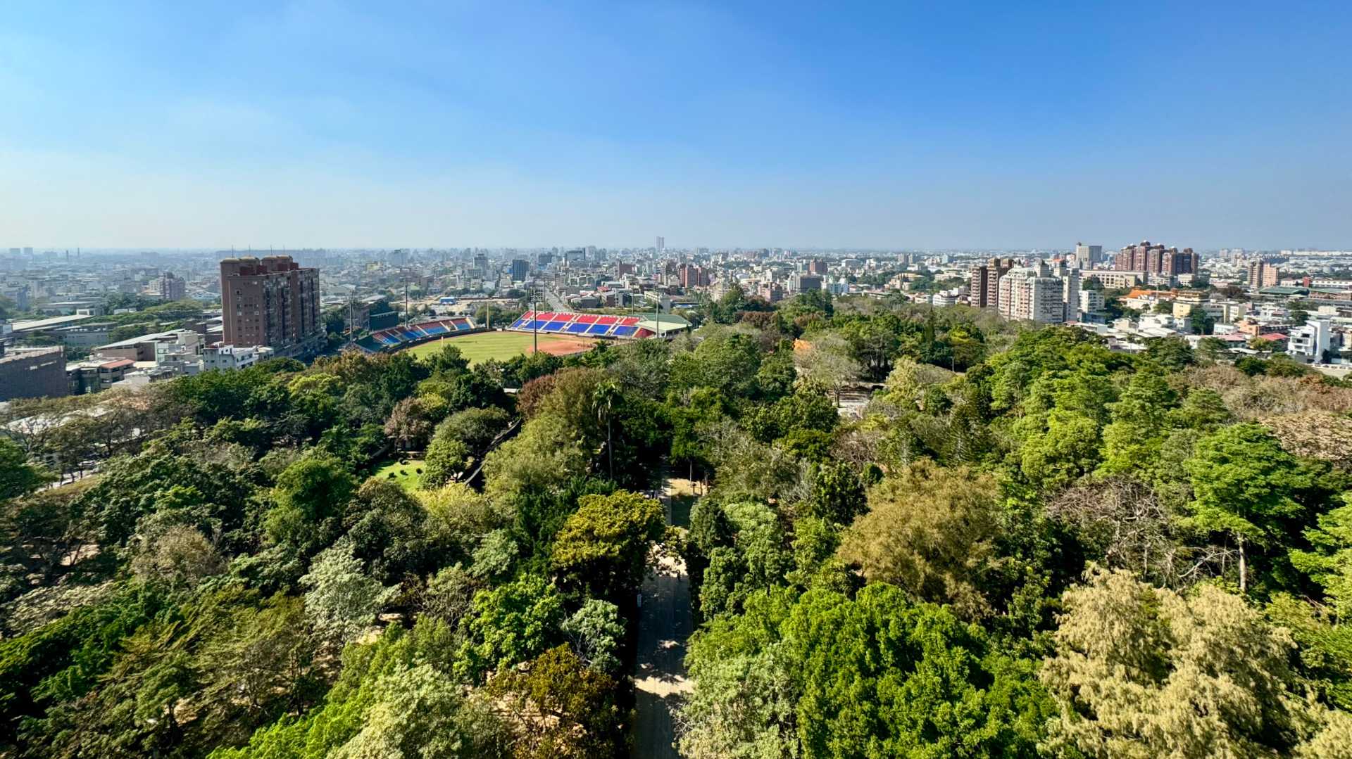 View across Chiayi City, Taiwan. The treetops of Chiayi Park are in the foreground. In the distance is a sports stadium, high rise buildings, and more buildings leading to the horizon.