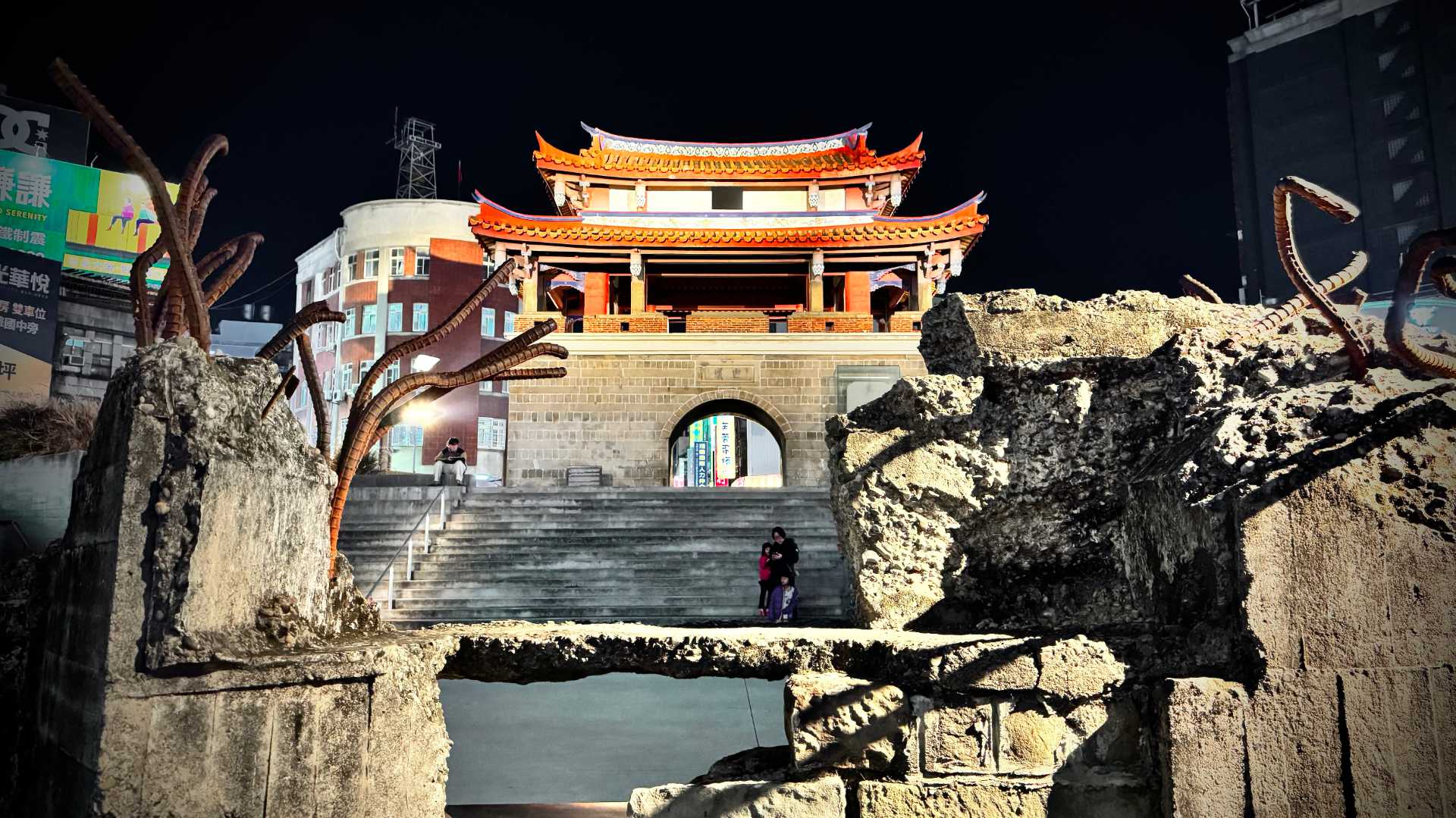 Night-time photo of Yieng-Siyi Gate in Hsinchu, a historic Qing Dynasty-era city gate, viewed through the ruins of a reinforced-concrete wall in the foreground.