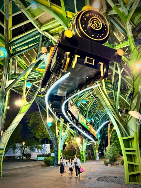 An large-scale public art installation of a half-real-size steam engine pulling four carriages full of cartoonish figures. The train is suspended from giant forest-like steel structures and appears to be flying on illuminated rails. About 5 meters below the train, three people are walking through this space carrying shopping bags.