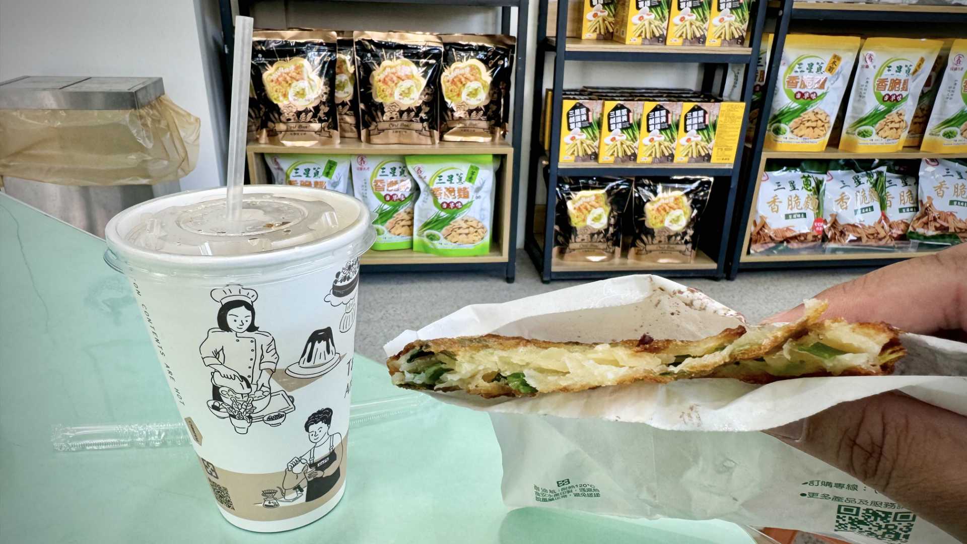 A take-out cup on a table, and a hand holding a paper bag that contains a partially-eaten scallion pancake.