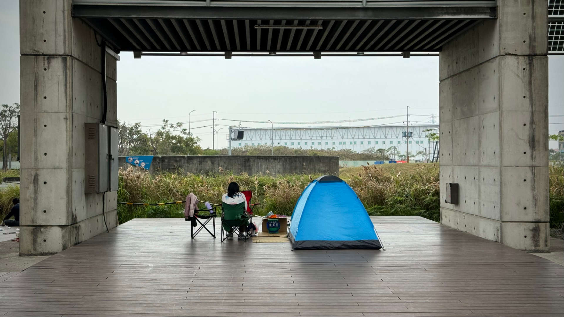 A lady in a camping chair, back to the camera, next to a small dome tent pitched under a solar array outside the National Museum of Taiwan History.