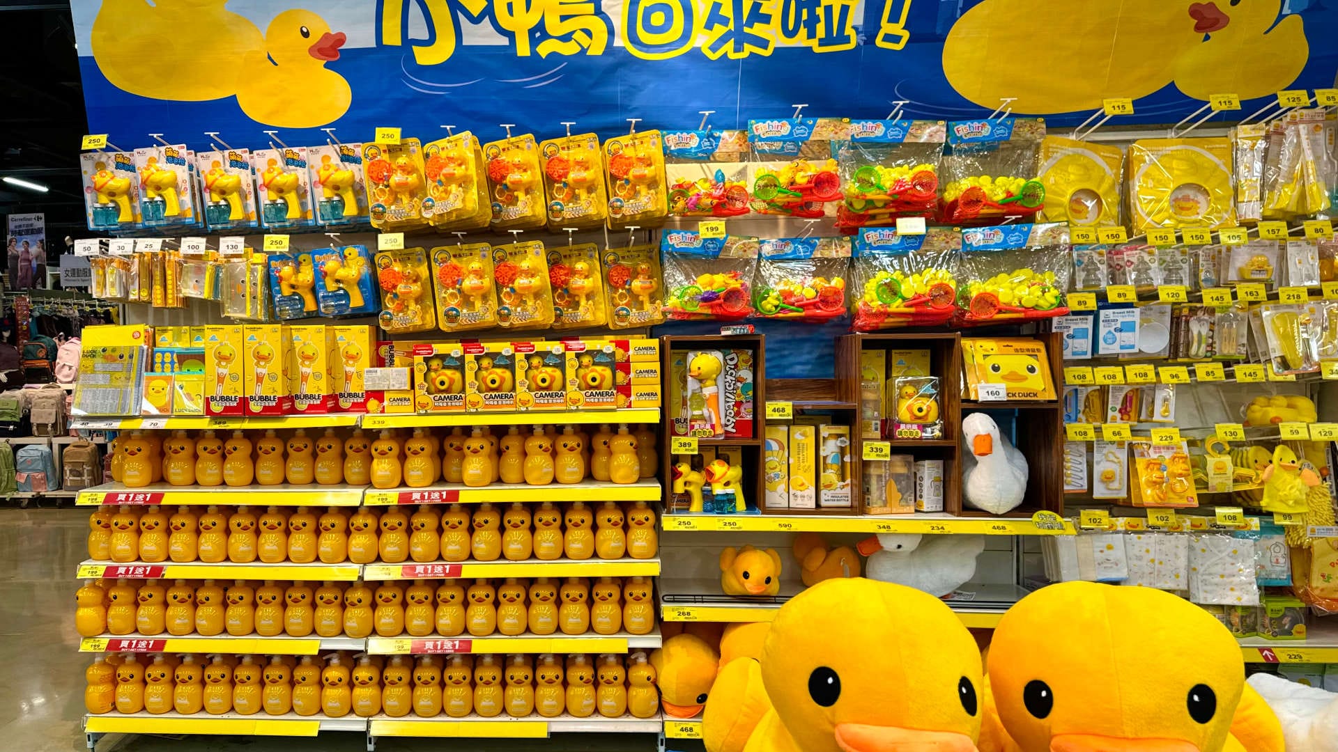 Supermarket shelves covered in yellow duck-themed items, such as liquid soap dispensers, bubble machines, cameras, rubber slippers, and carry bags.