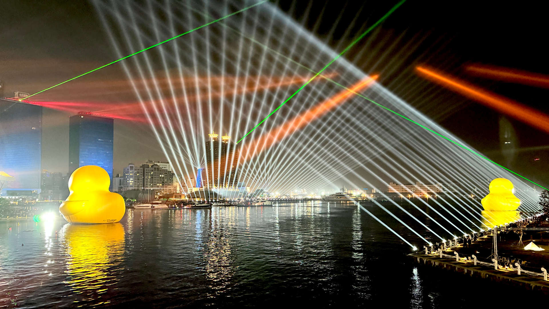 Night view of two large rubber ducks floating on Kaohsiung Harbor with a laser light show projecting dozens of multicolor beams of light into the dark sky.