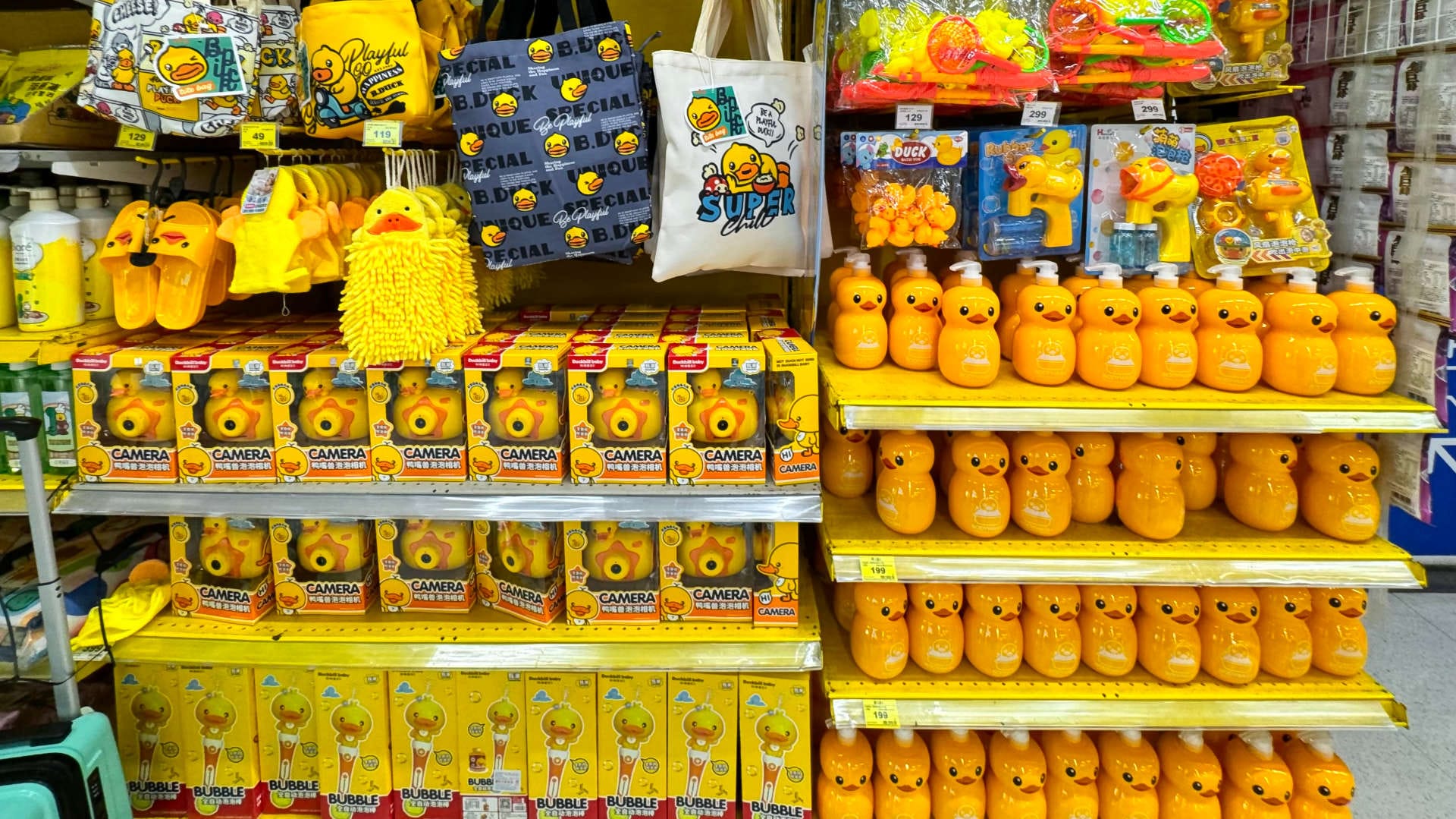 Supermarket shelves covered in yellow duck-themed items, such as liquid soap dispensers, bubble machines, cameras, rubber slippers, and carry bags.