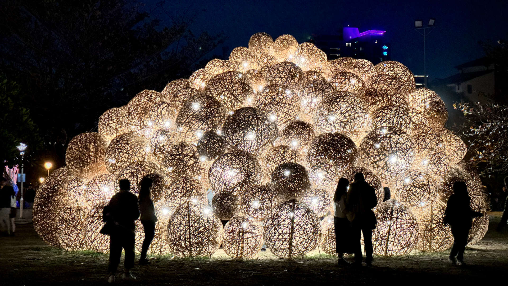 People are silhouetted in front of an illuminated sculpture made of dozens or hundreds of spheres. The spheres are made of curved wicker or some similar material.