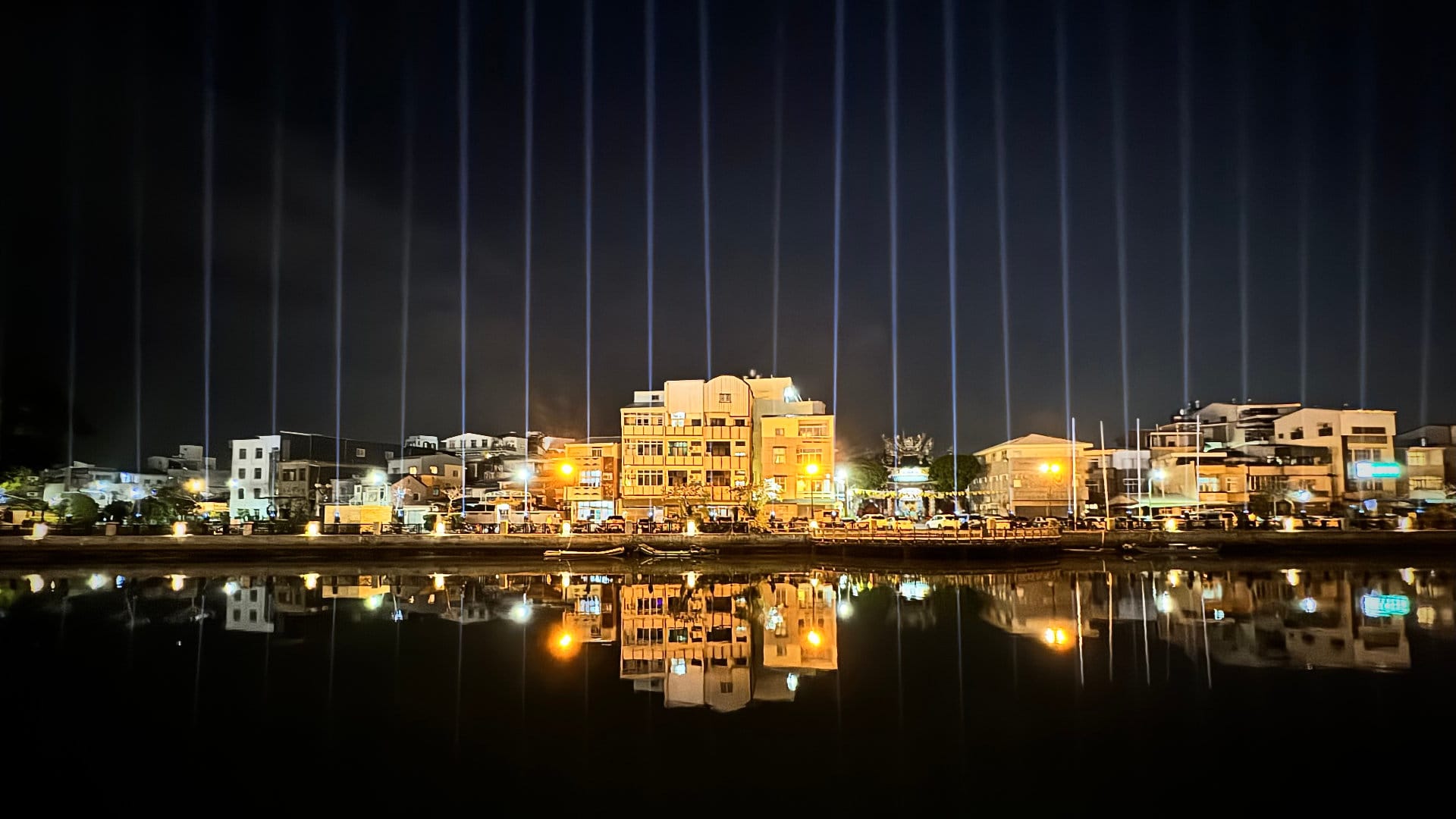 Looking across Tainan Canal. There are three-to-four-story buildings lining the canal. In front of them are dozens of parallel spotlights shooting parallel beams of light into the sky.