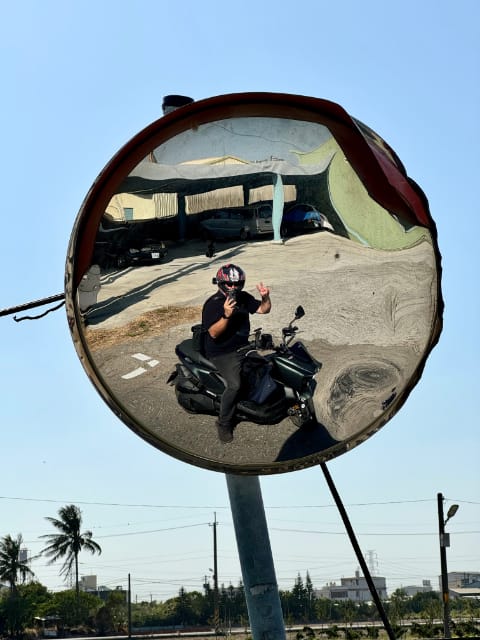 A distorted selfie taken in a convex roadside mirror. The photographer is sitting on his motorcycle and giving a peace sign with one hand.