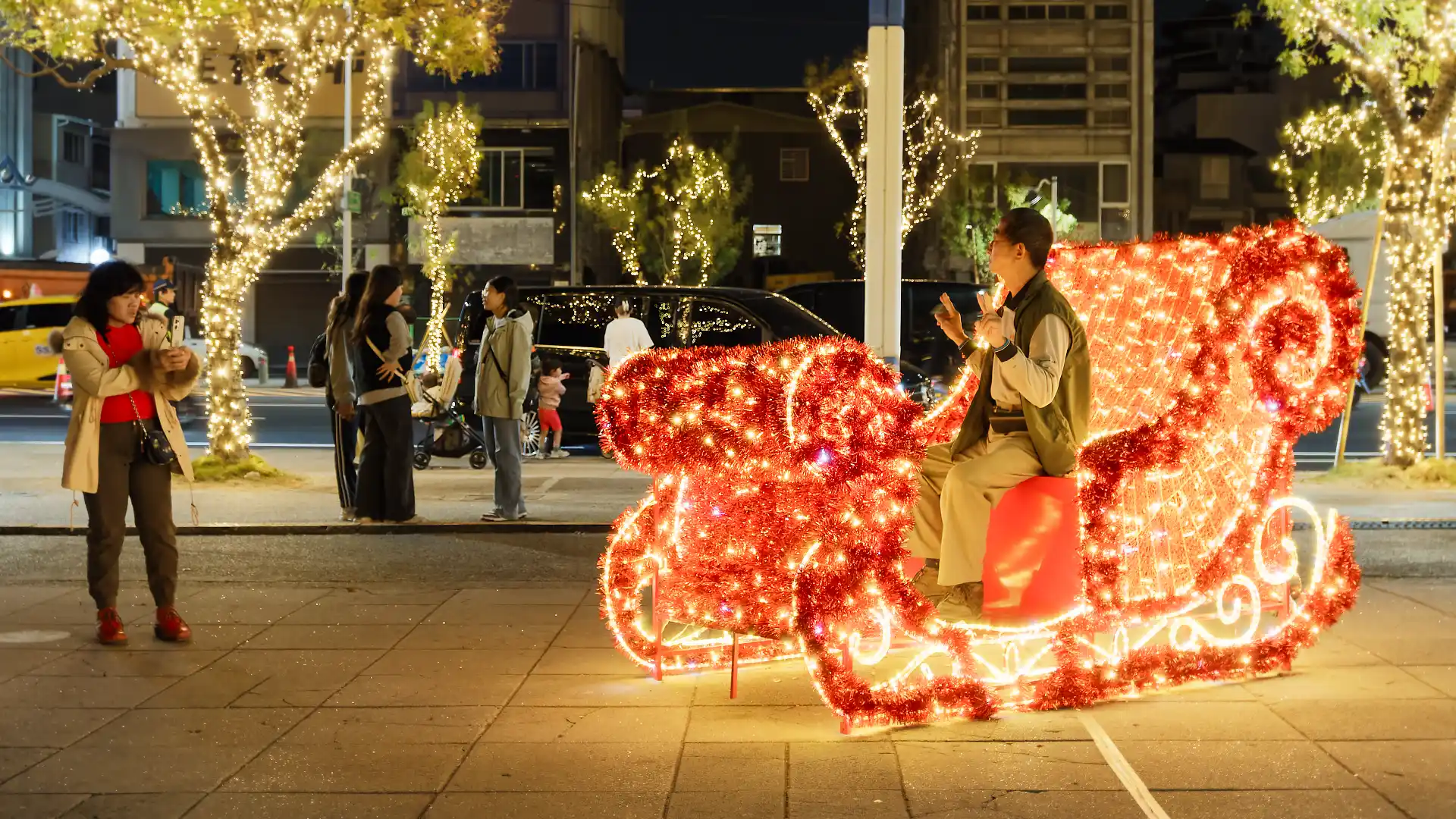A man poses for a photo while sitting in a sleigh made of red tinsel and string lights.
