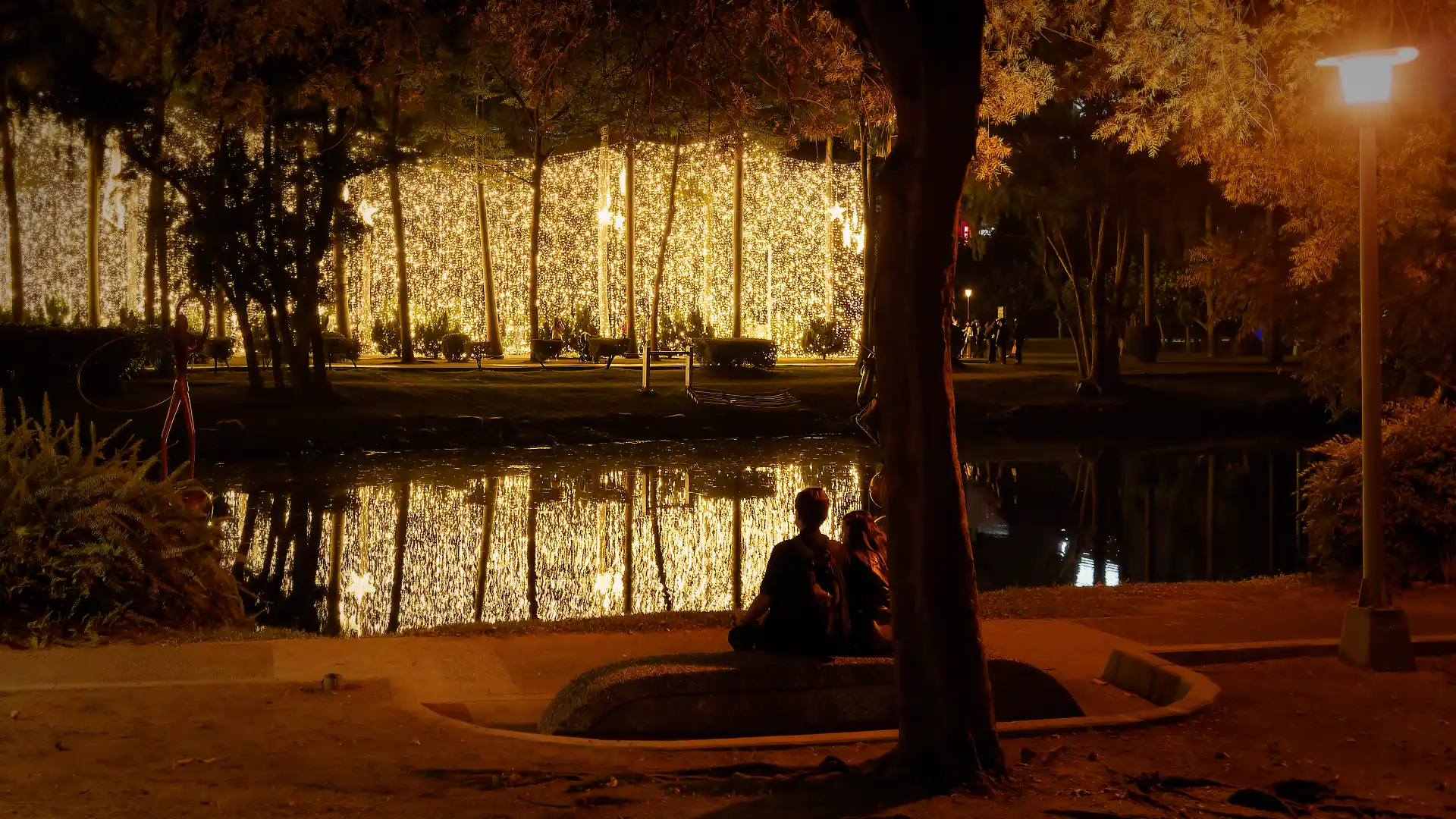 A couple silhouetted against the reflection of string lights in a still pond at Central Park, Kaohsiung, Taiwan.