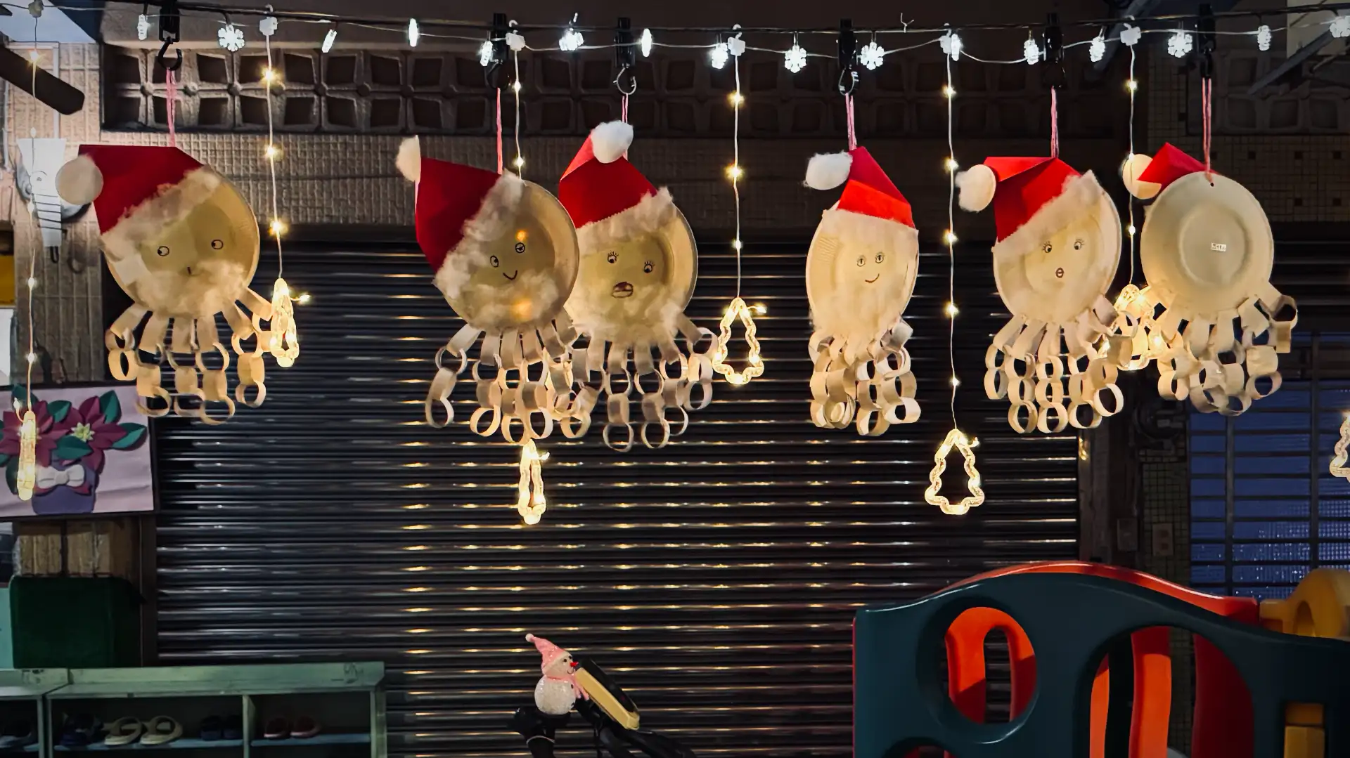 Six Santa decorations made of paper plates, hanging from a shop verandah and illuminated with Christmas lights.