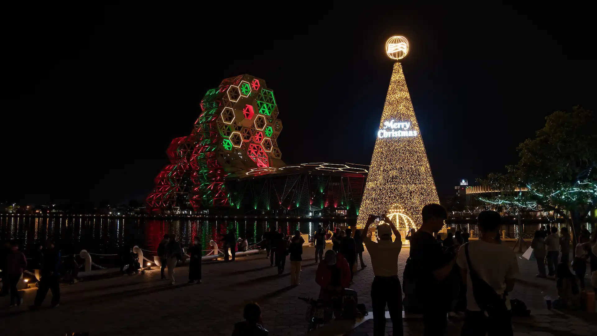An abstract cone-shape representing a Christmas tree, with Kaohsiung Music Center illuminated in red and green on the car side of Love River.