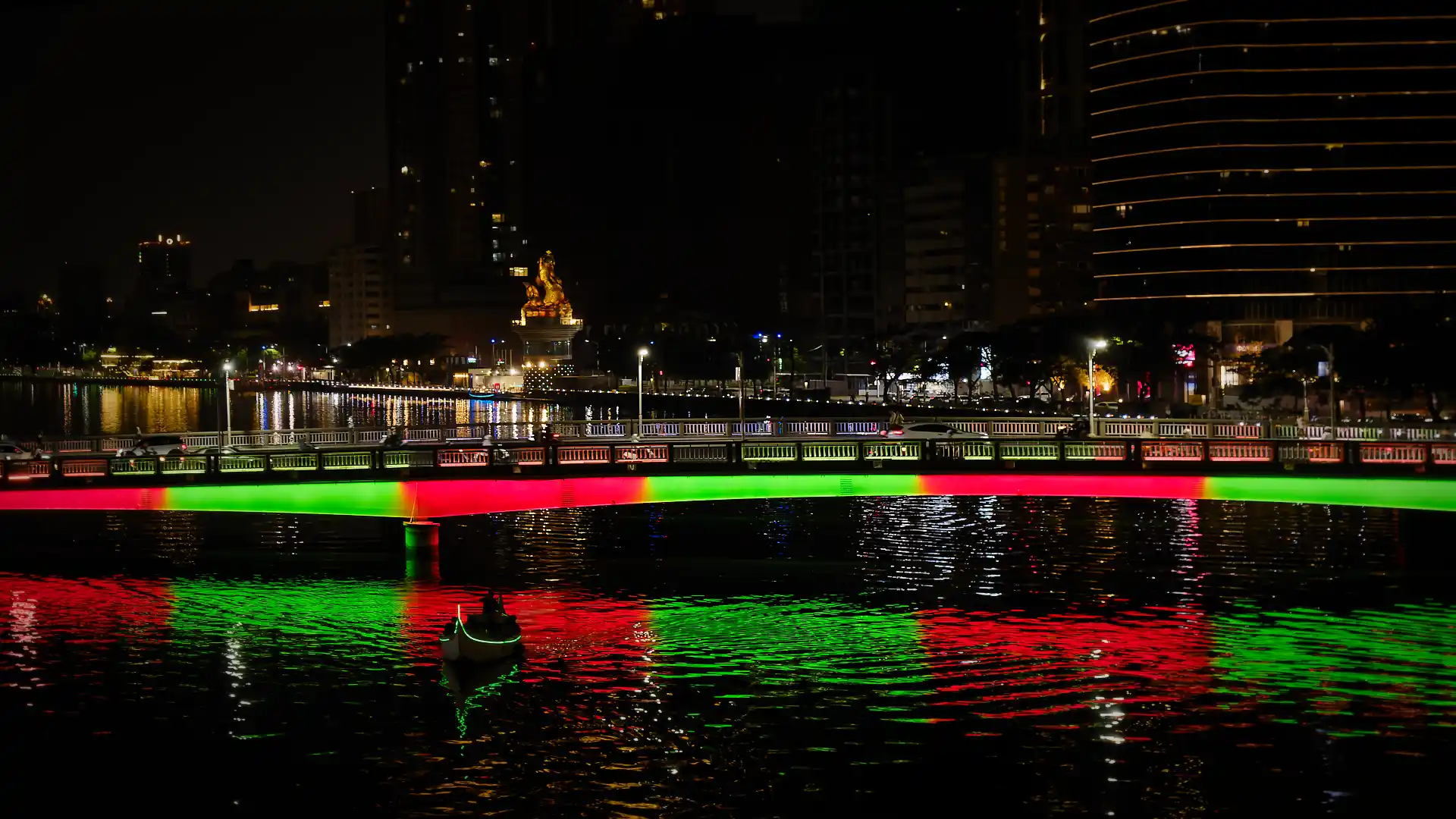A bridge over Love River is illuminated in red and green. The lights reflect in the river, where a Venetian-style gondola is silhouetted against the reflection.