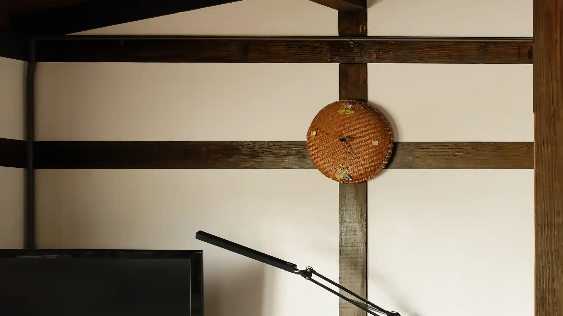 A wall clock of woven appearance above a TV and desk lamp.