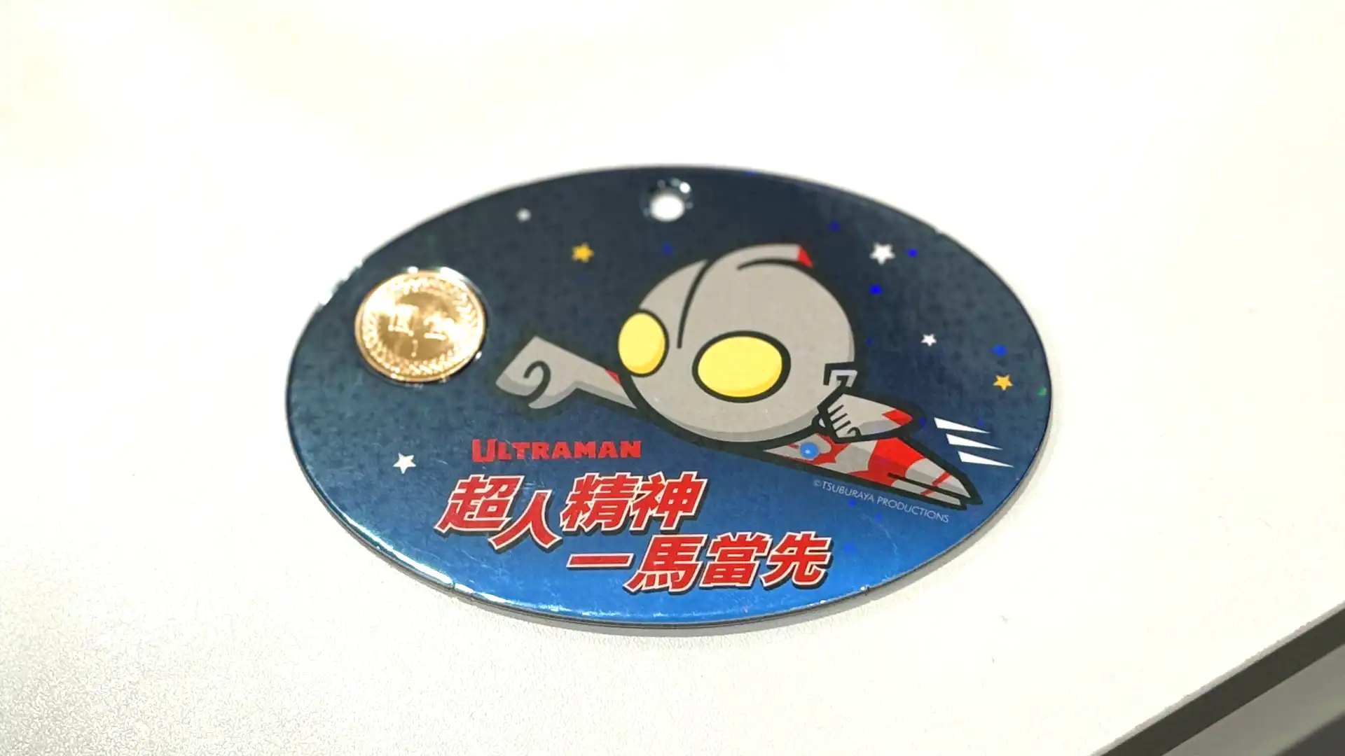 An oval-shaped thick collectable card featuring a cartoon image of Ultraman flying through space, and some Traditional Chinese writing.