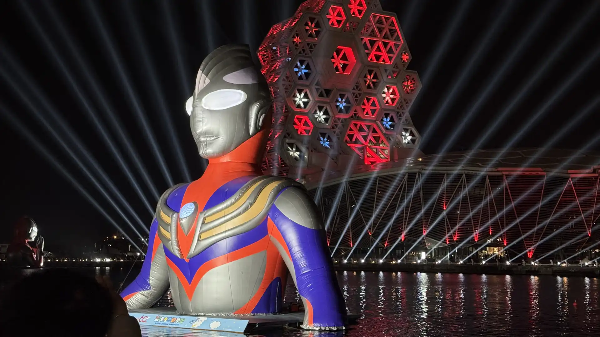 A giant inflatable Ultraman figure floating (waist-up) in Kaohsiung Harbor at night, with Kaohsiung Music Center lit up in special red lighting behind him. A couple of hundred meters away is a similar floating Ultraman figure in the distance.
