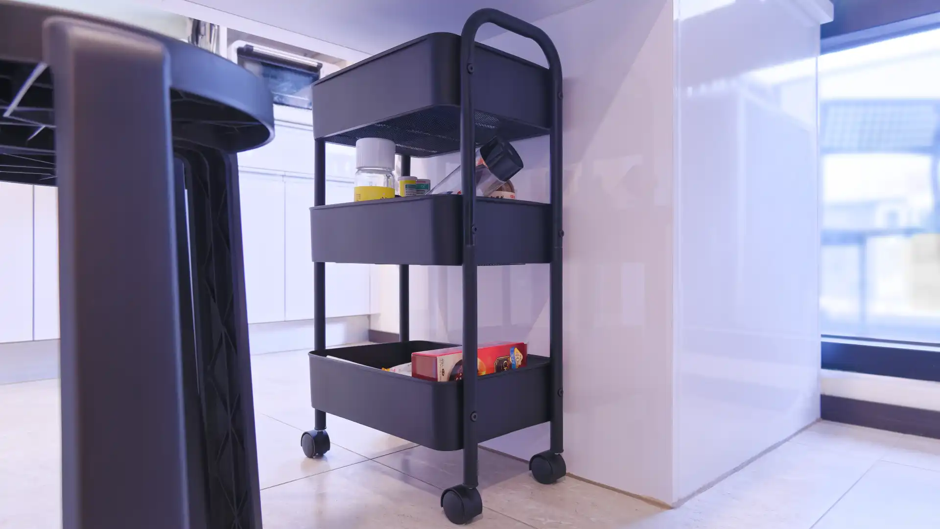 A small metal kitchen trolley wheeled under a kitchen counter and containing packets of processed foods.