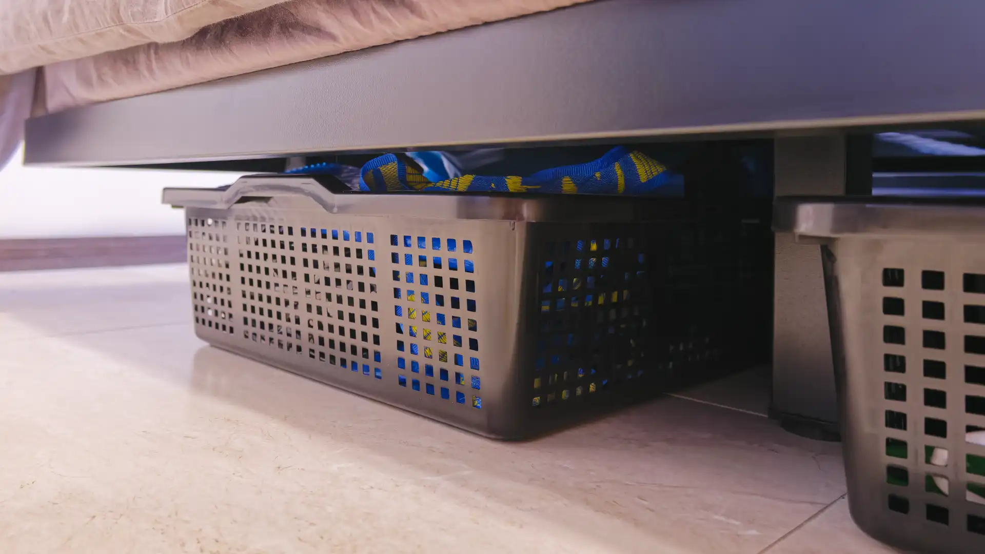 Plastic storage bins underneath a black metal bed frame. The baskets contain random household items including IKEA bags.