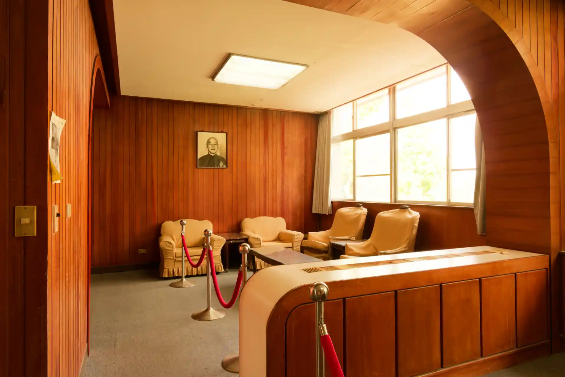 In the foreground is an inbuilt wooden storage unit, separating an office desk area from a sitting area. The sitting area has four lounge chairs arranged around a coffee table. A black and white portrait of Chiang Kai-Shek hangs on one wall.
