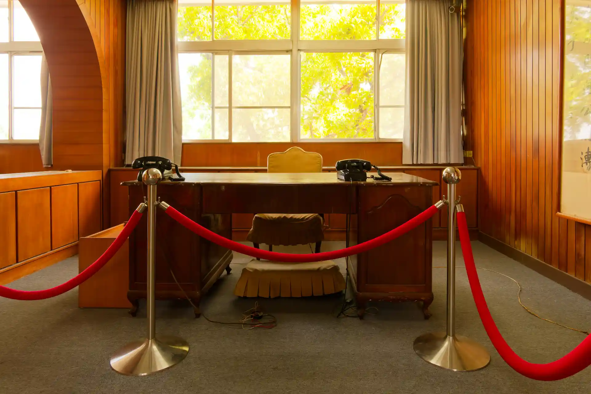 Beyond a velvet rope, an old wooden desk. Behind it is one velvet chair. On the desk are two black telephones.