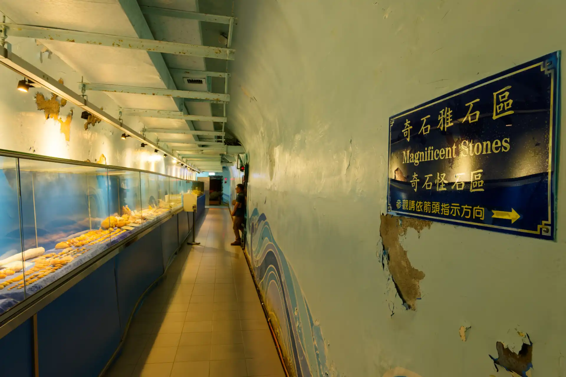 A long concrete corridor in a bunker, with illuminated display cabinets down the left wall. A sign on the right wall says “Magnificent Stones”.
