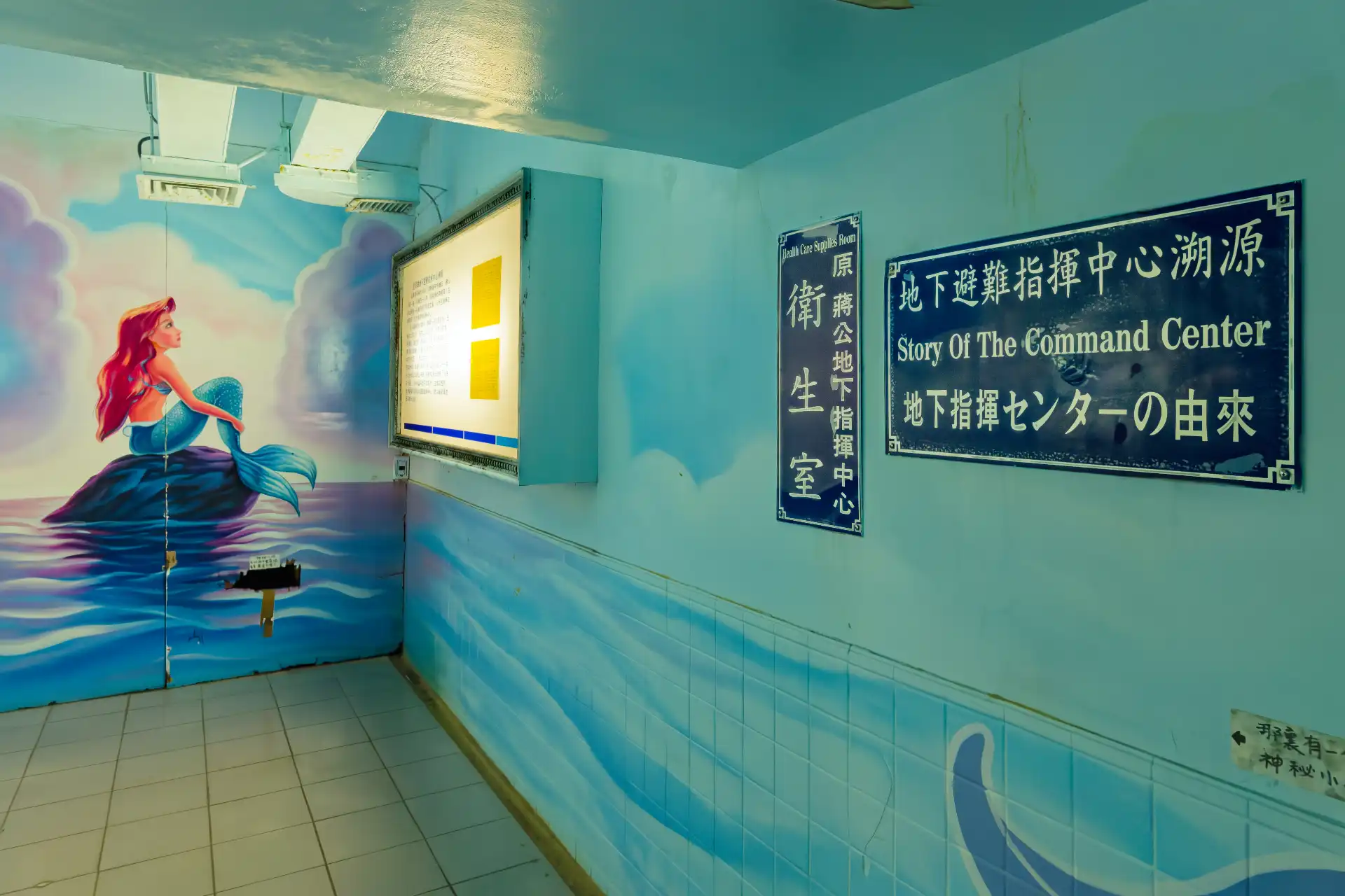 A square concrete room, decorated with a mermaid mural and a sign that says “Story Of The Command Center”. An illuminated light box at the far end of the room includes some text that’s too far away to read.