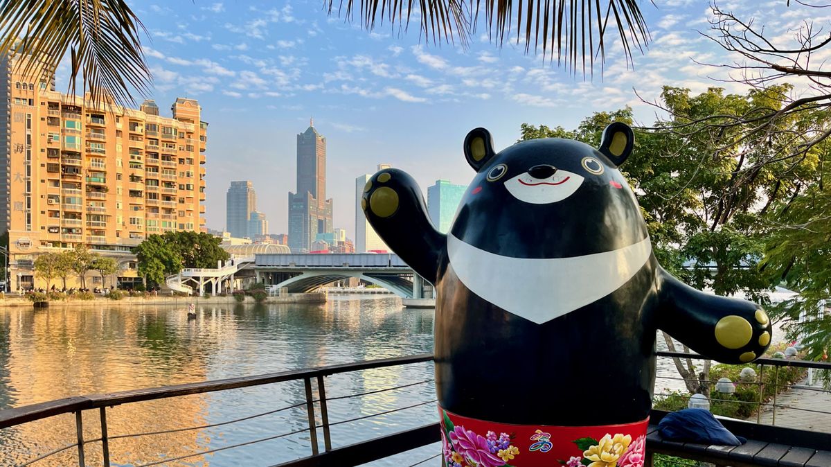 A human-size Formosan black bear mascot waving at the camera, with Love River and 85 Sky Tower in the background.
