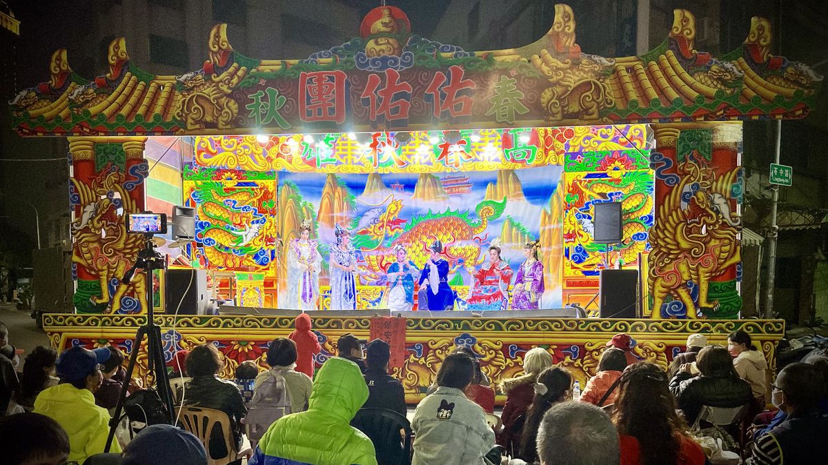 A temporary stage with six actors in traditional Chinese dress, watched by a crowd of people spread across the intersection.