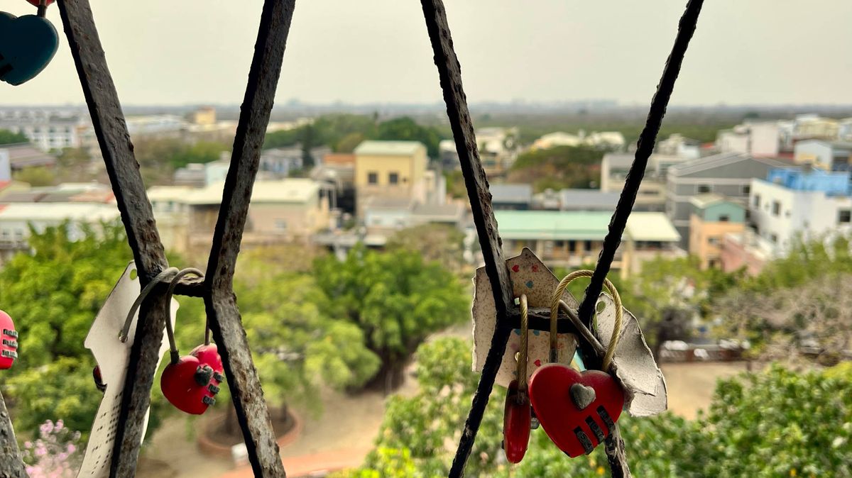 Heart-shaped padlocks attached to rusty metal bars, with a city view in the distance.