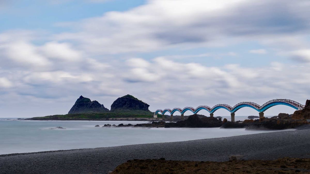 Long exposure of an eight-arched footbridge leading to a small forest-and-mountain-covered island.